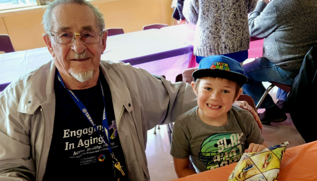 Older man and boy enjoy muffins at school hosted event Description: Seniors and elementary students connected in-person at an intergenerational event to have fun.