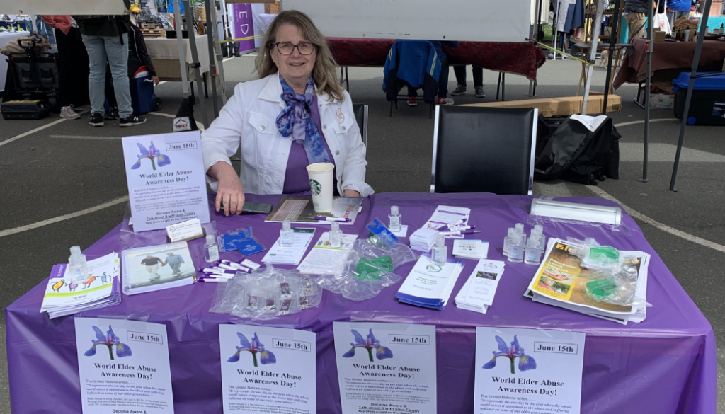 BC CRN Coordinator Gabi sits an an information table for WEAAD decorated in purple