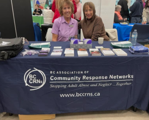 Two women smiling at the BC CRN information and resource table draped in a dark blue tablecloth